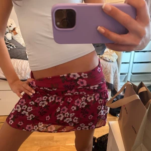 Vinröd  kortkjol - Säljer en röd kortkjol med blommigt mönster i rosa, vitt och grönt. Kjolen har en figurnära passform och är perfekt för dig som gillar färg och mönster. Superfin att styla med en enkel topp.