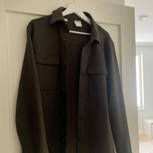 Mörkgrön overshirt från Selected Homme - Stilren mörkgrön overshirt från Selected Homme i storlek M. Skjortan har två bröstfickor med lock och knappar samt klassisk krage. Perfekt att bära som lager över en t-shirt. Materialet känns mjukt och slitstarkt. Änvand bara par gånger.