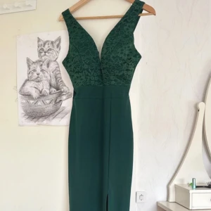 Grön klänning med spets och slits - Snygg grön klänning med djup v-ringning och blommig spets upptill. Klänningen har breda axelband, figurnära passform och en slits framtill. Tillverkad i polyester och passar perfekt för festliga tillfällen.