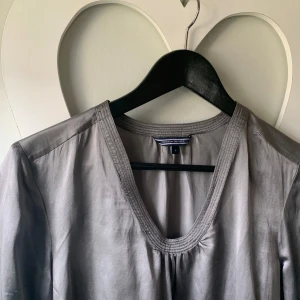 Grå långärmad blus från Tommy Hilfiger - Säljer en stilren grå långärmad blus från Tommy Hilfiger. Blusen har v-ringning med dekorativa sömmar runt halsen och är tillverkad i ett mjukt, lätt material som ger en snygg passform.