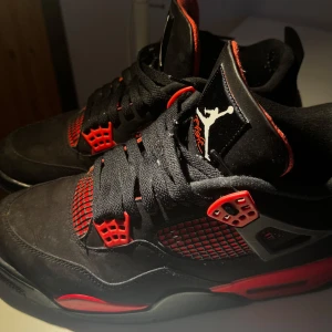Red Thunder Nike Air Jordans 4 - Säljer ett par Nike Air Jordans 4 i svart och rött med klassisk Jumpman-logga på hälen. Skorna har svarta snören, röda detaljer och meshpaneler på sidorna. Insidan är fodrad i rött tyg och sulan är röd med svarta inslag. Perfekta för dig som gillar streetwear. Orginal skolåda ingår!