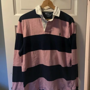 Randig rugbytröja från Polo Ralph Lauren - Säljer en klassisk rugbytröja från Polo Ralph Lauren i rosa och marinblå breda ränder. Tröjan har vit krage, lång ärm och broderad grön logga på bröstet. Perfekt för en sportig och stilren look.