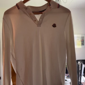 Vit pikétröja från Moncler - Säljer en klassisk vit pikétröja från Moncler med lång ärm och knappar vid halsen. Tröjan har Moncler-logga på bröstet och krage med rödblå randig detalj. Köpt för cirka 3000 på NK (inget kvitto) NFC Scan funkar och qr kod.
