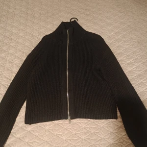 Svart ribbad kofta med dragkedja - Säljer en svart ribbad kofta med hög krage och dragkedja framtill. Tröjan är croppad och har långa ärmar, perfekt att slänga på sig över en t-shirt eller linne.