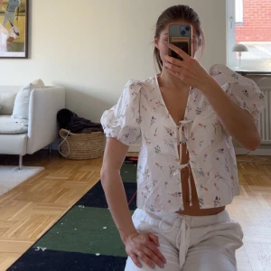 Vit blommig blus med puffärmar - Söt vit blus med små blommor i olika färger. Blusen har puffiga korta ärmar, knytning framtill och volangdetaljer vid ärmslut. Perfekt för en somrig look och ger en romantisk känsla. 