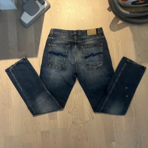 Blå jeans från Nudie Jeans Co - Säljer ett par klassiska blå jeans från Nudie Jeans Co i grim tim modellen med raka ben och snygga kontrastsömmar. Jeansen har normal passform och ikonisk broderad bakficka. Perfekta för dig som gillar stilrena och tidlösa jeans.      Det finns 2 små fläckar på det högra benet av jeansen som helt ärligt ser bara ut som designen😊