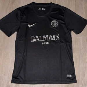 Svart PSG x Balmain fotbollströja - Säljer en svart PSG x Balmain fotbollströja från Nike. Tröjan har korta ärmar, rund hals och vita detaljer med klubbmärke och Nike-logga på bröstet samt Balmain Paris-tryck på framsidan. Tillverkad i lätt och ventilerande material.
