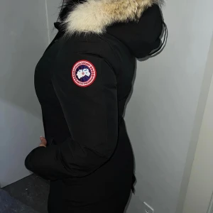 Svart parkajacka med päls från Canada Goose - Säljer en svart parkajacka från Canada Goose med avtagbar huva och äkta päls runt kragen. Jackan har knappar framtill, två fickor och klassisk logotyp på ärmen. Perfekt för kalla dagar.Har även kvar lappen. Pris kan diskuteras