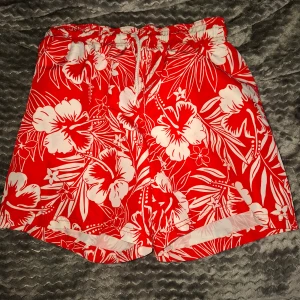 Röda blommiga badshorts - Snygga röda badshorts från Name It med vitt blommigt mönster och elastisk midja med snörning. Perfekta för sommardagar vid stranden eller poolen.