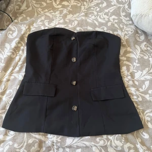 Svart tubtopp med knappar - Snygg svart tubtopp med knappar framtill och två dekorativa fickor. Toppen har en stilren och elegant design, perfekt för att lyfta vilken outfit som helst. Gjord i ett slätt material som ger en dressad känsla.