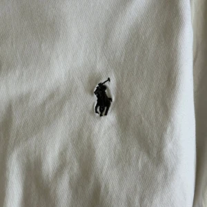 Vit t-shirt från Polo Ralph Lauren - Klassisk vit t-shirt från Polo Ralph Lauren med broderad svart logga på bröstet. T-shirten har rund hals och korta ärmar. Perfekt basic-plagg med stilren design och skön passform.