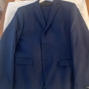 Mörkblå kostym från Jack & Jones - Säljer en stilren mörkblå kostym från Jack & Jones i modellen Super Slim, storlek 50. Kostymen består av en kavaj och matchande byxor ( storlek 52), perfekt för bal eller fest. Klassisk design med knappar och fickor på kavajen.