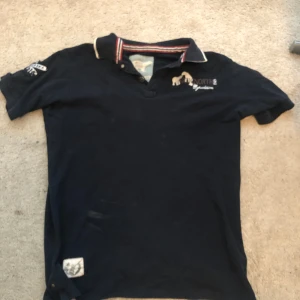 Mörkblå pikétröja från Artic North - Mörkblå pikétröja med broderade isbjörnar och flaggor på ryggen samt logga på bröstet. Kragen har rödvita detaljer.