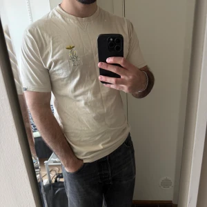 Beige t-shirt med broderad blomma - Säljer en beige t-shirt med en liten broderad blomma och text på bröstet. T-shirten har rund halsringning och korta ärmar. Perfekt för en avslappnad stil.