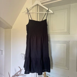 Svart sommarklänning med smock och knytband - Säljer en perfekt svart sommarklänning från Cotton On med smock upptill och tunna axelband. Klänningen har knytband i ryggen och volang nertill. Perfekt för varma dagar och enkel att styla med sneakers eller sandaler💗