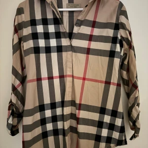 Burberry skjorta strl M - Klassisk skjorta från Burberry i beige med stort rutmönster i svart, vitt och rött. Skjortan har lång ärm, knappar framtill och en tidlös design. Perfekt för dig som gillar ikoniska mönster och stilren look. Storlek M. Skjortan behöver en tvätt. Garanterar att det är äkta Burberry!