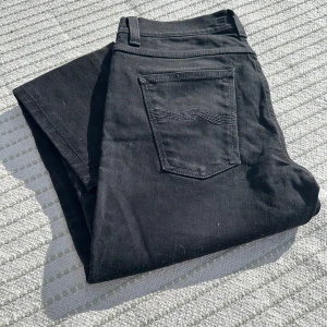 Svarta jeansbyxor från Nudie Jeans - Säljer ett par svarta jeans från Nudie Jeans med klassisk femficksdesign och raka ben. Byxorna har svarta sömmar och en diskret logga på bakfickan. Perfekta för dig som gillar stilrena och tidlösa jeans. Skick 9/10 W30L30