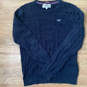 Mörkblå stickad tröja från Hampton Republic - Mörkblå stickad tröja från Hampton Republic med diskret logga broderad på bröstet. Tröjan har ett klassiskt kabelstickat mönster och rund halsringning. Perfekt för lager-på-lager och passar till många olika stilar.