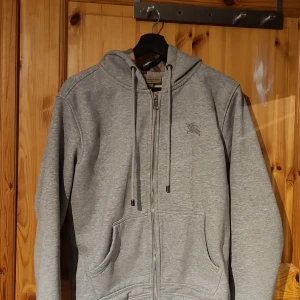 Grå hoodie från Burberry - Säljer en klassisk grå hoodie från Burberry med dragkedja och fickor framtill. Huvan har det ikoniska rutmönstret på insidan. Perfekt för en avslappnad stil och passar året runt. Fråga gärna om fler bilder och priset kan diskuteras vid snabb affär.