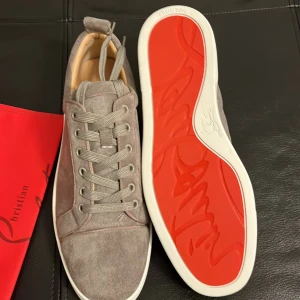 Grå sneakers från Christian Louboutin - Säljer ett par gråa sneakers i mocka från Christian Louboutin med klassisk röd sula. Skorna har rund tå, snörning och vit platt sula. Perfekta för dig som gillar stilrena och exklusiva sneakers. Priset kan sänkas vid snabb och smidig affär!!