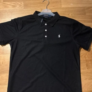 Svart pikétröja från Polo Ralph Lauren - Helt ny, klassisk svart pikétröja från Polo Ralph Lauren med vit broderad logga på bröstet. Tröjan har krage, tre knappar framtill och är kortärmad. Perfekt för en stilren och tidlös look.