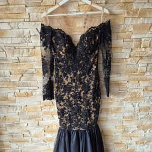 Svart långklänning med spets och lite paljetter - Säljer en elegant svart långklänning med transparent mesh och vackra spetsdetaljer samt paljetter. Klänningen har offshoulder-modell, lång ärm och figurnära passform. Knäppning med knappar i ryggen.
