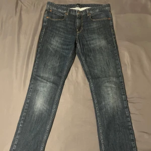 Mörkblå jeans från Hugo Boss - Säljer ett par klassiska mörkblå jeans från Hugo Boss med raka ben och snygga slitningar. Jeansen har fem fickor, orange sömmar och läderpatch med logga bak i midjan. Perfekta för en stilren look.
