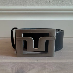 Svart skärp från J. Lindberg - Säljer ett svart bälte från Lindberg med ett stort, fyrkantigt silverfärgat spänne. Bältet är i skinn och har en stilren design som passar till många olika outfits.