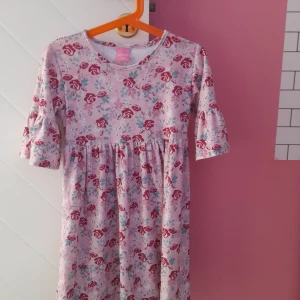 Rosa blommig klänning med volangärm - Söt rosa klänning med blommigt mönster i rött och blått. Klänningen har rund halsringning och volangdetaljer på de halvlånga ärmarna. Perfekt för vår och sommar!