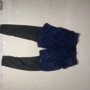 Blå träningsbyxor med svarta tights - Säljer ett par blå träningsbyxor med inbyggda svarta tights. Byxorna har resår i midjan och dragsko samt fickor på sidorna. Perfekta för löpning eller gym. Sportig look och bekväm passform.
