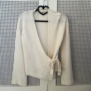 Vit omlottkofta med knyt - Säljer en mjuk och mysig vit omlottkofta från ginatricot. Använd en gång.  Koftan har lång ärm, v-ringad hals och knyt i sidan för en snygg passform. Perfekt att slänga på sig när det är lite kyligt ute. Materialet är stickat och känns riktigt skönt mot huden.