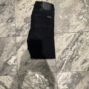 Svarta jeans från Nudie Jeans, 25/32 - Snygga svarta jeans från Nudie Jeans i modellen 25/32. De har klassisk femficksdesign, svarta sömmar och en stilren läderpatch bak i midjan. Jeansen är raka och har en smal passform som sitter riktigt bra. Perfekta för dig som gillar minimalistisk stil.