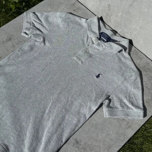 Grå polo t-shirt från Ralph Lauren - Snygg grå polo t-shirt från Ralph Lauren i slim fit. Klassisk krage, två knappar och broderad logga på bröstet. Tillverkad i mjuk bomull som är skön mot huden. Perfekt för dig som gillar stilrena och tidlösa plagg.