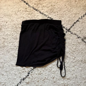 Svart kortkjol med snörning - Säljer en svart kortkjol med snygg snörning på sidan som ger en cool, asymmetrisk look. Kjolen är ribbad och har en elastisk midja för skön passform. Perfekt för dig som gillar stilrena plagg med en twist.