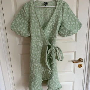 Grön blommig omlottklänning  - Supersöt ljusgrön omlottklänning från Pieces i storlek XS. Klänningen har puffiga korta ärmar, knytband i midjan och ett vitt blommigt mönster. V-ringad och med volangkant nedtill, perfekt för vår och sommar. Materialet känns lätt och luftigt.