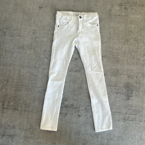 Name It vita byxor  - Snygga vita byxor från Name It i barnstorlek 128. Klassisk femficksmodell med dragkedja och knapp framtill. Byxorna har smal passform.