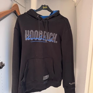 Svart hoodie från Hoodrich med blått tryck - Svart hoodie från Hoodrich med stor logotyp i grått och blått på bröstet och ryggen. Tröjan har huva med blått foder, snörning och en stor magficka. Långärmad och tillverkad i mjuk bomull, perfekt för chill dagar. Snygga detaljer med broderad text på ärmen.
