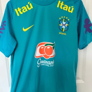 Blå Brasilien fotbollströja Nike - Brasilianska landslagets träningströja med CBF-emblem och flera sponsorer som Itaú och Guaraná Antarctica. Tröjan har korta ärmar, gul Nike-logga och lila tryck på ärmarna. Tillverkad i lätt och ventilerande polyester, perfekt för träning eller match. 