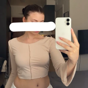 Beige topp - Säljer en beige croppad topp med långärmade, lätt utsvängda ärmar och en synlig söm framtill. Toppen har en rund halsringning och är tillverkad i ett ribbat material som ger en snygg och modern look.