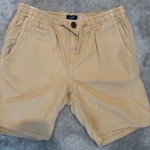 Beige shorts med snörning från Superdry -  Superdry Shorts ✨ Snygga beige shorts från Superdry 🤎 med elastisk midja och snörning 👌. 	•	👖 Två sidofickor och bakfickor med knapp framtill 	•	🌸 Insidan har ett coolt blommigt mönster i marinblått 	•	🌞 Perfekta för avslappnade sommardagar  Bekväma och stilrena shorts för en chill och trendig look. 