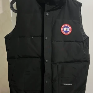 Svart dunväst från Canada Goose - Svart dunväst från Canada Goose med klassisk logga på bröstet. Västen har två stora fickor framtill, knäppning med tryckknappar och hög krage. Perfekt för lager-på-lager och riktigt snygg till streetwear eller outdoor-stil.