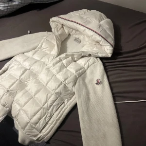 Vit quiltad pufferjacka från Moncler - Snygg vit pufferjacka från Moncler med quiltad front och huva med broderad logga. Ärmarna är stickade och har Moncler-märke på vänster arm. Jackan har dragkedja och en clean, modern look som passar perfekt till kalla dagar. Den har nfc tar byten beror på vad det är strl 1-s men passar även M mycket  pris kan diskuteras 