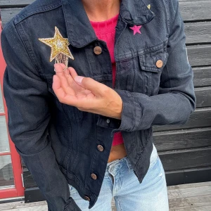 Svart jeansjacka blingad och dekorerad stl.XS - Cool svart jeansjacka i stl.XS från H&M med flera färgglada stjärnpatchar och en broderad 'oops!' på ryggen. Jackan har knappar framtill, klassisk krage och långärmade armar. Materialet är robust denim och detaljerna gör den unik och personlig.