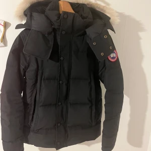 Svart dunjacka från Canada Goose - Säljer en svart dunjacka från Canada Goose med fluffig beige päls på huvan. Jackan har quiltad design, dragkedja och knappar framtill samt praktiska fickor med dragkedja. Perfekt för kalla vinterdagar och har den klassiska loggan på ärmen.