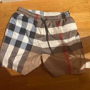 Rutig Burberry shorts  - Snygga Burberry shorts i klassiskt rutigt mönster med beige, blå, svart, vit och röd. Tillverkade i 100% bomull med meshfoder och elastisk midja med snörning. Perfekta för sommaren och ger en clean och lyxig vibe.