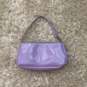 Ljuslila axelväska med glansig finish - Snygg ljuslila axelväska med glansig yta och stilren, avlång form. Väskan har ett smalt axelband och stängs med dragkedja upptill. Insidan är ljusbeige och rymmer det viktigaste. Perfekt för att addera färg till din outfit.
