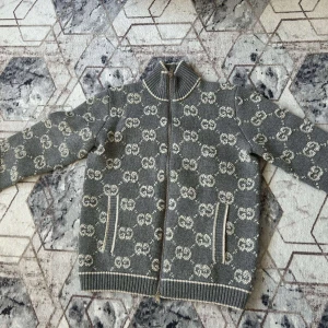 Grå stickad cardigan jacka från Gucci - Snygg grå stickad cardigan jacka från Gucci med hel dragkedja och ribbad krage. Jackan har ett mönster med vita GG-loggor över hela och två fickor framtill med vita detaljer. Perfekt för dig som gillar exklusiv streetstyle och vill sticka ut.