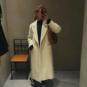 Vit kappa från Sandro Paris - Snygg benvit kappa från Sandro Paris med oversized passform, stora slag och dubbelknäppning med guldiga knappar. Jackan är lång, har fickor fram och är tillverkad i en ull, bomull och alpackamix som ger en mjuk och lyxig känsla.