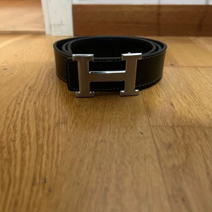 Svart skärp med H-spänne - Snyggt svart skärp med ett stort, silvrigt H-spänne i metall. Bältet är gjort av skinn och har vita sömmar längs kanterna som ger en clean kontrast. Perfekt för att lyfta din outfit med en lyxig detalj.