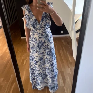 Blommig långklänning med volanger - Säljer en superfin långklänning med blått blommönster. Klänningen har  v-ringning, volangdetaljer vid axlarna och öppen rygg med snörning. Perfekt för sommarens och den har en flowy och luftig passform. Den är från hm i fint skick i storlek 36🥰